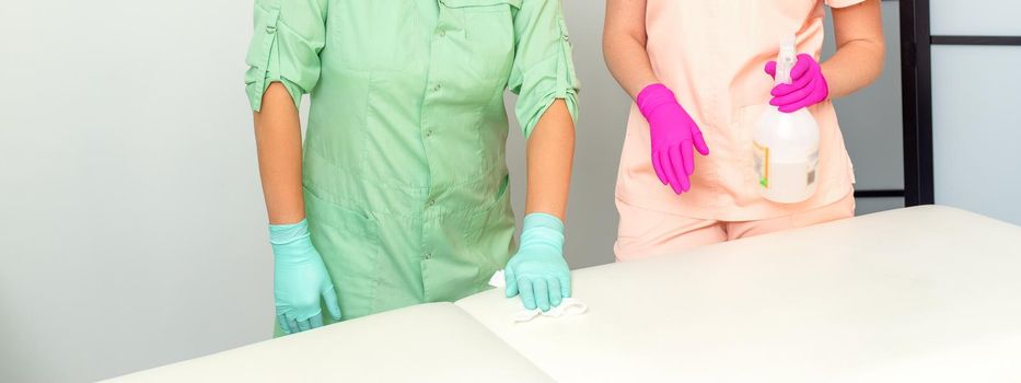 Two medical workers disinfect the patient's couch with sanitizer spray and a clean napkin. Health and hygiene concept.