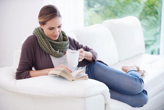 Lost in a page-turner...A young woman relaxing on her couch with a book and a cup of coffee.