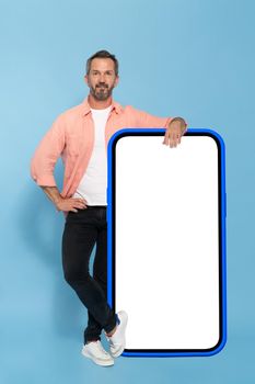 Middle aged fit man leaned on huge, big smartphone with white screen in blue case happy smiling on camera wearing peach shirt and black jeans isolated on blue background. Mobile app advertisement