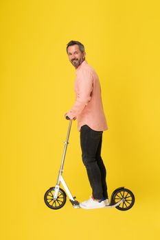 Middle aged grey haired man ride scooter standing sideways looking at camera wearing peach shirt and black jeans isolated on yellow background. Fit masculine handsome mature businessman on the go