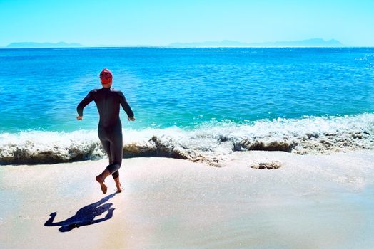 Endurance swimming. Rearview shot of a female swimmer in a wetsuit running towards the surf.