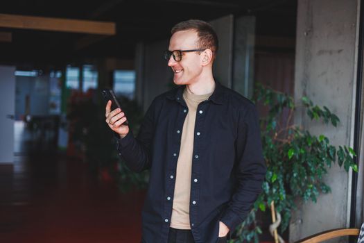 Half-length portrait of happy man in optical glasses holding modern mobile phone and smiling at camera, cheerful blogger with digital smartphone posing in coworking space