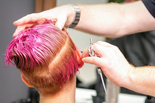 Haircut of dyed short pink wet hair of young caucasian woman by a male hairdresser in a barbershop.