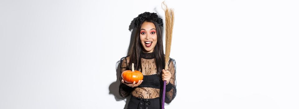 Excited attractive asian girl celebrating halloween, wearing witch costume, holding pumpkin and broom, going trick or treat, standing over white background