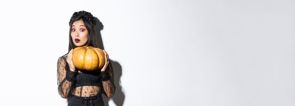 Portrait of woman lifting heavy pumpkin, getting ready for halloween, wearing witch costume, standing over white background