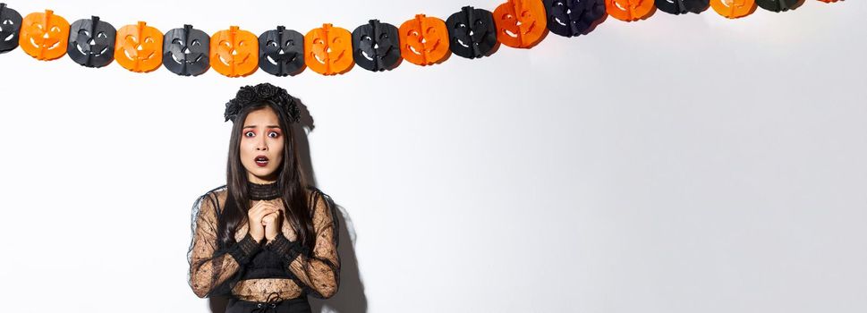 Image of scared and worried asian woman in witch costume looking concerned, standing against pumpkin streamers decoration