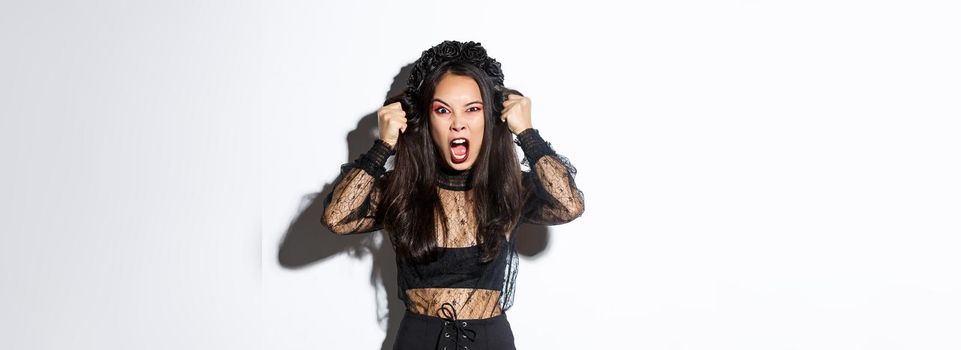 Angry asian woman impersonate evil witch on halloween party, looking mad and furious, screaming to scare people, standing over white background in gothic dress