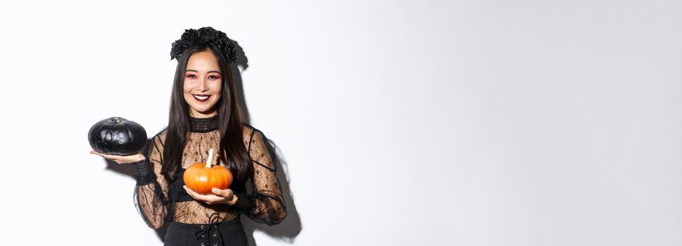 Image of attractive korean girl celebrating halloween in gothic lace dress, impersonating witch and holding two pumpkins