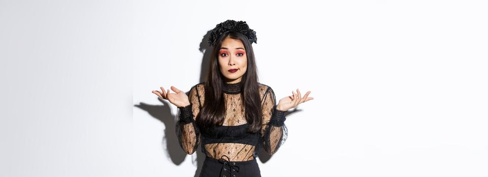 Image of clueless and unbothered asian girl looking confused, shrugging while standing over white background in halloween costume, dressed-up like evil witch on party