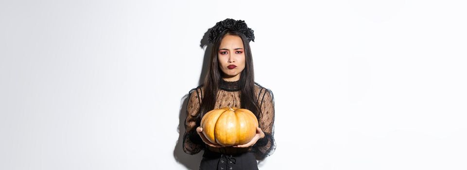 Image of grimacing asian woman in witch costume hate carve pumpkin for halloween, looking disappointed, standing over white background
