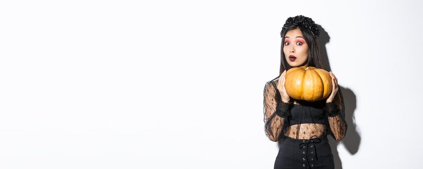 Portrait of woman lifting heavy pumpkin, getting ready for halloween, wearing witch costume, standing over white background