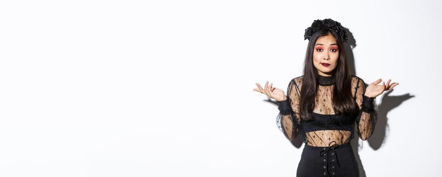 Image of clueless and unbothered asian girl looking confused, shrugging while standing over white background in halloween costume, dressed-up like evil witch on party