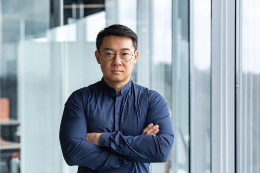 Portrait of successful and serious asian boss in shirt, man looking at camera with crossed arms inside modern office building near window