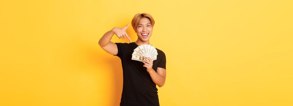 Sassy good-looking korean blond guy, smiling happy and pointing finger at money, winning cash, standing yellow background