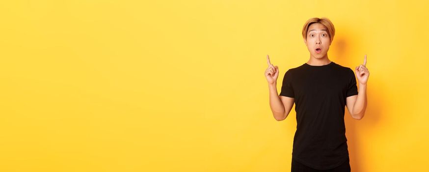 Portrait of astonished asian guy in black t-shirt, open mouth fascinated, pointing fingers up, yellow background