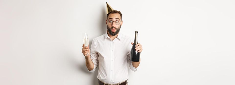Celebration and holidays. Surprised man wearing birthday hat, holding champagne and glass and looking amazed, standing over white background