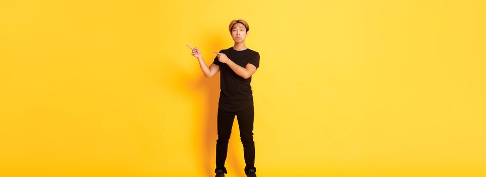 Full-length of sad and miserable asian guy in black clothes, pouting, pointing fingers upper right corner, standing yellow background