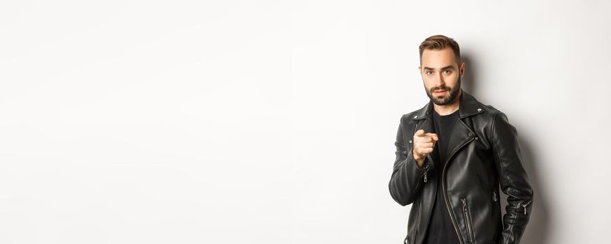 Handsome and cool bearded guy pointing finger at camera, wearing leather jacket, standing sassy against white background