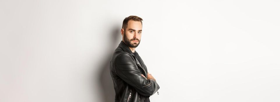Profile of confident and handsome bearded man in black leather jacket, turn face at camera and looking serious, white background
