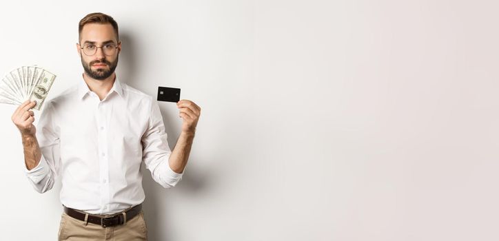 Serious businessman looking at camera, holding credit card and money, standing over white background. Concept of shopping and finance