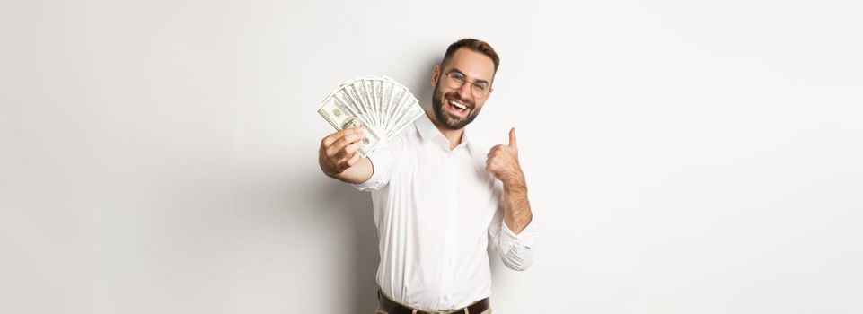 Successful businessman showing money dollars and thumbs-up, smiling satisfied, standing over white background