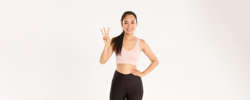 Sport, wellbeing and active lifestyle concept. Smiling sportswoman, asian fitness girl showing three useful exercises for losing weight or gaining muscles, standing white background in activewear