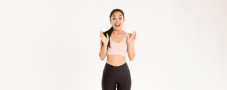 Sport, wellbeing and active lifestyle concept. Happy enthusiastic fitness girl, asian sportswoman clasp hands from fantastic news, gasping amazed, standing over white background