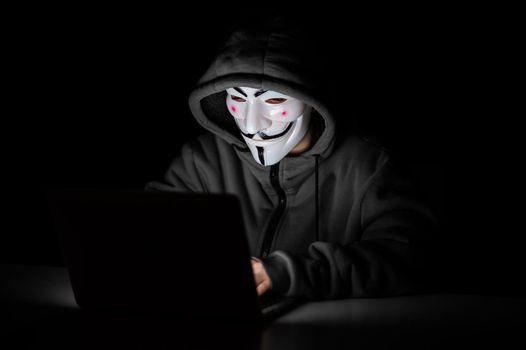 Anonymous in a hood is typing on a laptop in the dark.