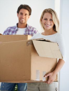 Moving into a home of her own. Portrait of a smiling woman carrying a brown cardboard box with her boyfriend carrying a box in the background.