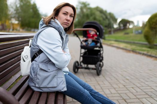 annoyed young mother sitting in the park on a bench away from the baby stroller with an angry face. the concept of postpartum depression