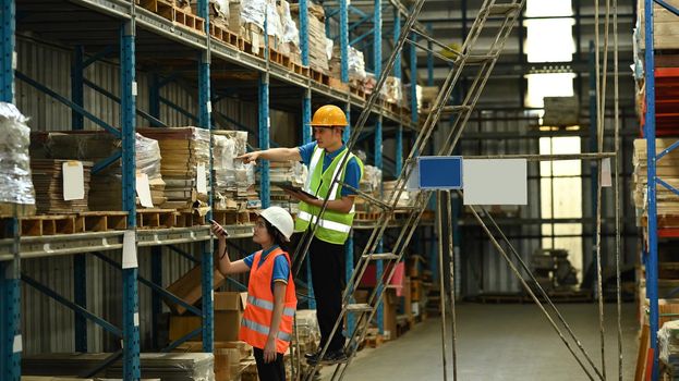 Male and female workers wearing hardhats and reflective jackets checking inventory boxes on shelf with barcode scanner
