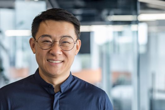 Close up photo portrait of successful young Asian investor, man in glasses smiling and looking at camera in shirt, businessman working inside modern office building, satisfied accountant financier