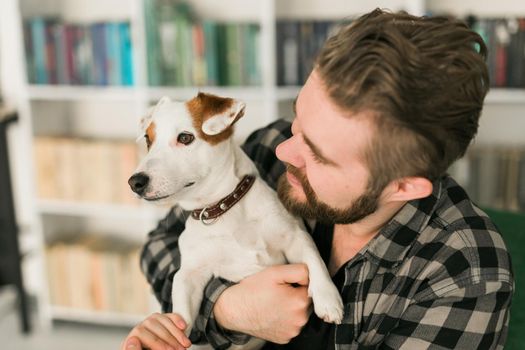 Happy male owner of jack russell terrier dog, feels responsibility of caring about pet, standing against bookshelves background. People and relationship with animals.