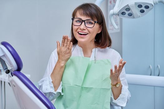 Happy middle aged woman patient looking at camera sitting in dental chair in dentist office. Smiling female after taking care of teeth whitening treatment, dental implant prosthetics