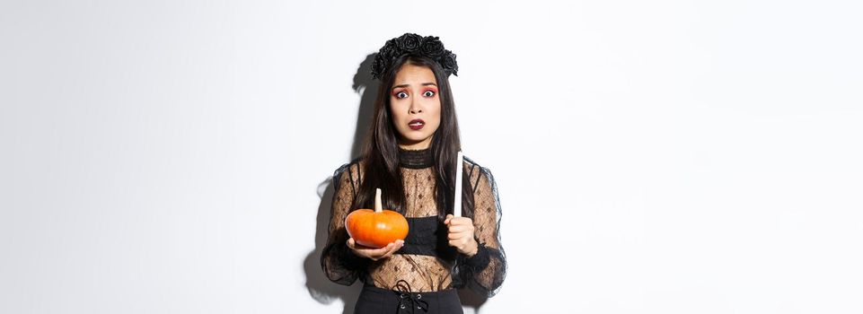Image of asian woman panicking, being scared of witchcraft on halloween, standing in witch costume with candle and pumpkin