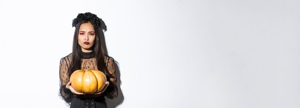Image of grimacing asian woman in witch costume hate carve pumpkin for halloween, looking disappointed, standing over white background