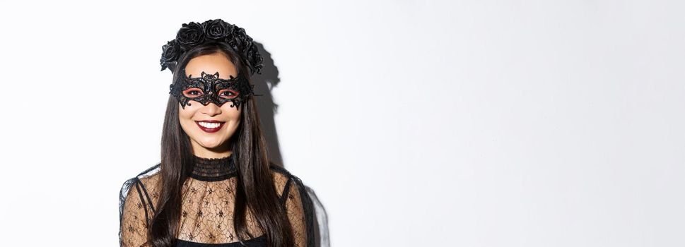 Close-up of mysterious woman in gothic wreath and black mask smiling at camera, celebrating halloween, standing over white background