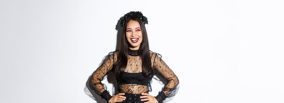 Image of carefree smiling asian woman in gothic dress and black wreath looking thoughtful at upper left corner, licking lips from desire or temptation, standing over white background