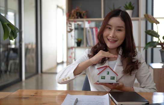 Miniature house in the hands of an Asian woman real estate agent home loan working at the office. Looking at the camera.