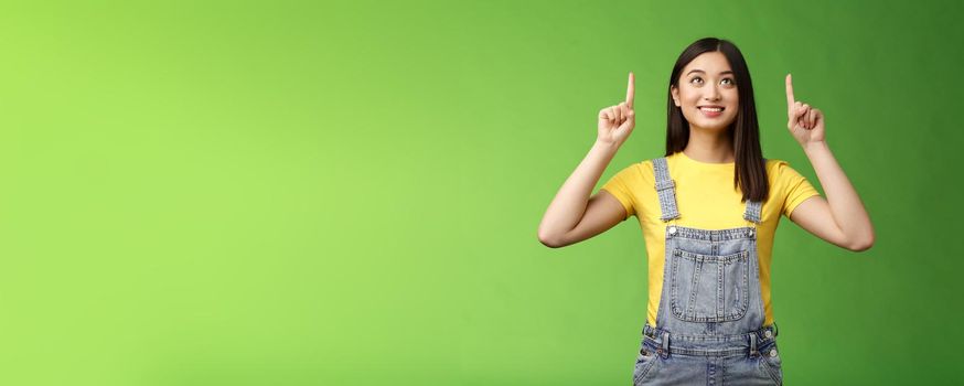 Dreamy hopeful cute asian girl look pointing up amused, smiling delighted, contemplate interesting object, enjoy stargazing, grinning satisfied, gazing top promo, stand green background