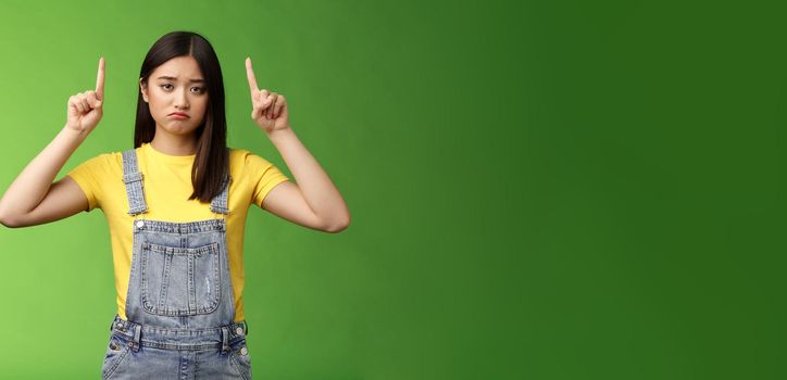 Pity cute sad asian girl brunette, grimacing pulling sorrow upset face frowning bothered, pointing fingers up regret awful situation, indicate top promo, stand green background unhappy
