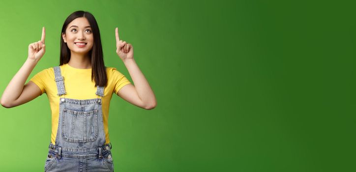 Dreamy hopeful cute asian girl look pointing up amused, smiling delighted, contemplate interesting object, enjoy stargazing, grinning satisfied, gazing top promo, stand green background