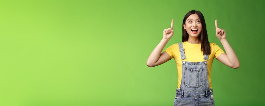 Carefree amused asian brunette in yellow t-shirt dungarees, smiling amazed and fascinated, look pointing up, observe cool new promo, grinning excited, enjoy watching top copy space