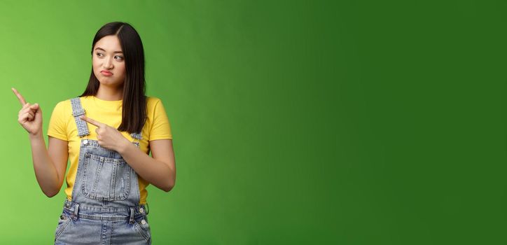 Upset cute asian female student pulling face regret shame, pity missing good chance buy product sale, pointing looking left disappointed, jealously gaze coworker, stand green background
