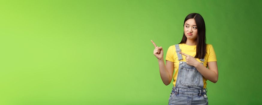 Upset cute asian female student pulling face regret shame, pity missing good chance buy product sale, pointing looking left disappointed, jealously gaze coworker, stand green background