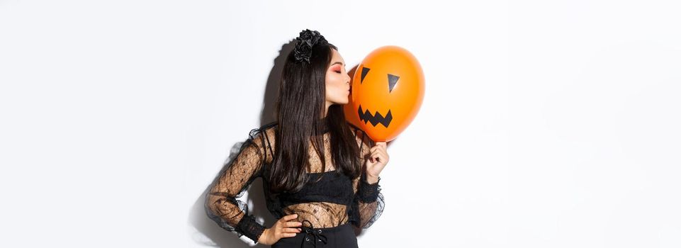 Stylish asian woman in gothic lace dress celebrating halloween, kissing orange balloon with face, standing over white background
