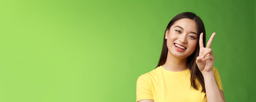 Close-up joyful cute asian girl lively look camera smile, show peace victory sign entertained, grinning posing photograph tender expression, stand green background positive and lucky