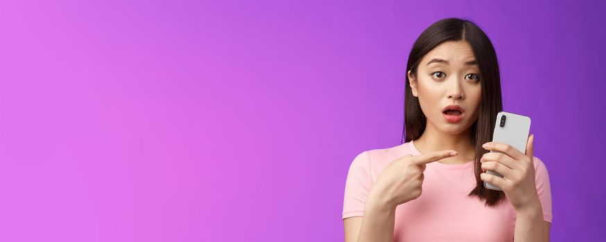 Concerned confused cute young asian woman hold smartphone open mouth, pointing finger telephone, look questioned upset, troubled see strange post online, stand purple background
