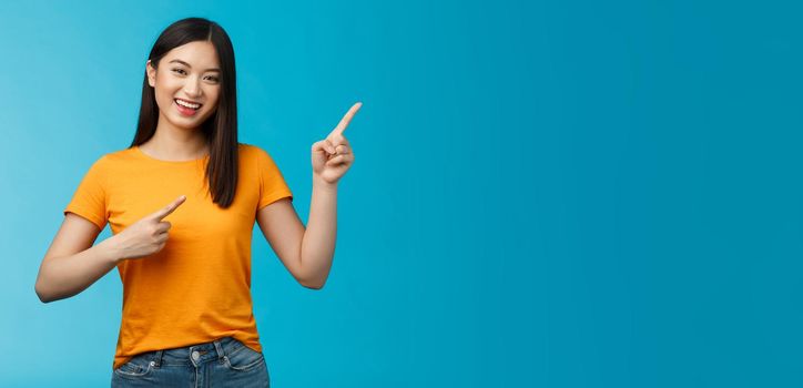 Cheerful lively cute asian woman smiling laughing upbeat, pointing upper right corner, give advice what choose, shopping online, grinning happily, enthusiastic promo, blue background