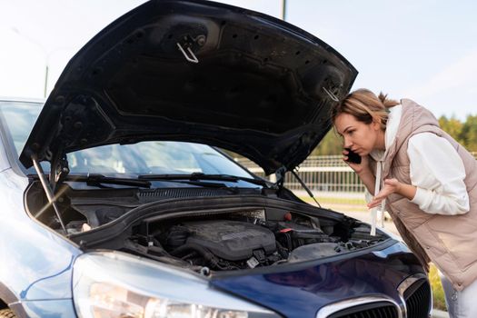 confused female driver looking under the hood talking on a mobile phone and trying to understand the cause of the car breakdown